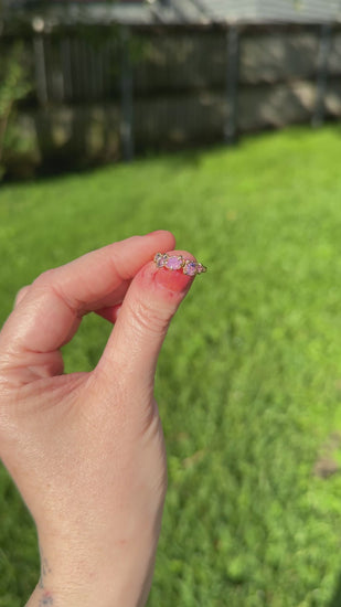Pink and purple Freeform sapphire engagement ring in woman’s fingers with sparkly pink nail polish with grass in the background